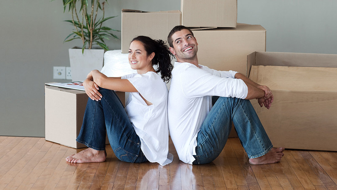 Glückliches Paar sitzt Rücken an Rücken auf einem Holzparkett-Fußboden vor ihren Umzugskartons.