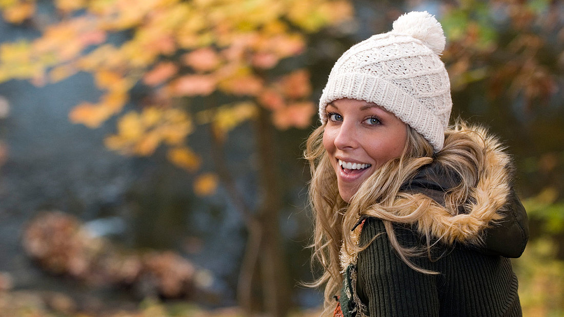 Junge, blonde Frau läuft warm verpackt mit Mütze, Schal und dicker Winterjacke glücklich durch eine Herbstlandschaft.