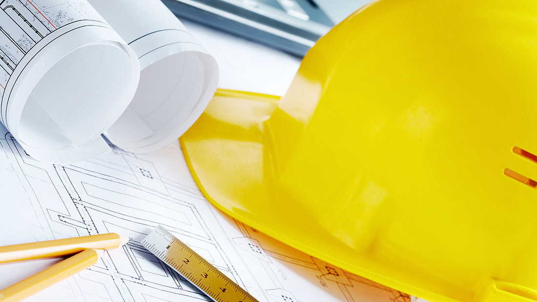 Utensilien eines Bauunternehmens ausgebreitet auf einem Tisch, z.B. Bleistifte, Bauplan und Bauhelm.